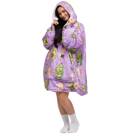 Eine Person trägt einen übergroßen Avokado Familie Hoti-Kapuzenpullover in Lila mit Avocado- und Toastmustern der Marke The Hoti. Sie hat lange Haare und posiert mit einem verspielten Lächeln, die Hände vor dem Gesicht. Vor dem schlichten weißen Hintergrund sind weiße Socken zu sehen.