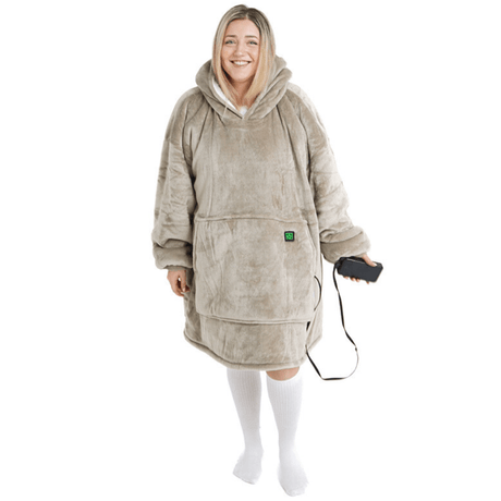 Eine Person lächelt, während sie den Flauschiger Beige Hoti von The Hoti trägt, eine übergroße Kapuzendecke aus weichem grauem Material mit einer großen Vordertasche. Sie hält ein kabelgebundenes Gerät in der Hand und trägt lange weiße Socken.