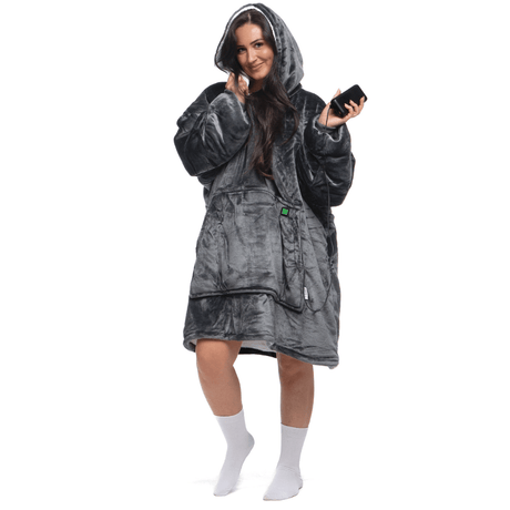 Eine Person, eingehüllt in einen bequemen Kapuzenmantel „Grauer Zauber Hoti“ von The Hoti, hält ein Smartphone. Sie steht vor einem weißen Hintergrund, lächelt und trägt weiße Socken.